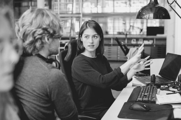 Software engineer sitting at her desk, explaining something to her colleague with expressive hands.